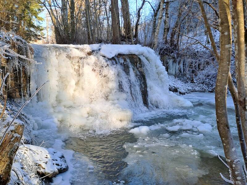 Eine verschneite Winterlandschaft zeigt einen teils zugefrorenen Wasserfall, umgeben von schneebedeckten Bäumen.