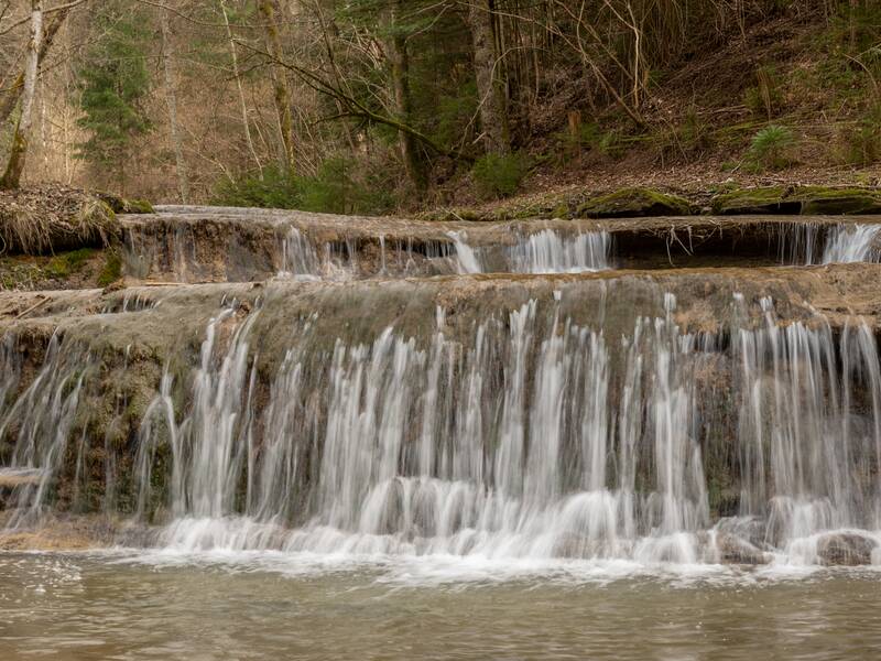 Ein kleiner Wasserfall fließt über mehrere Stufen eines steinigen Flussbetts in einem bewaldeten Gebiet.