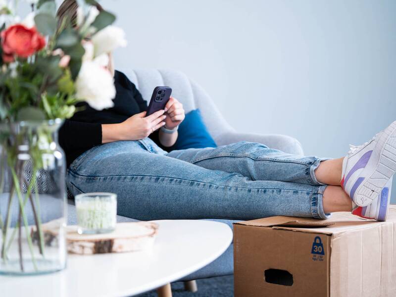 Eine Person mit einem Smartphone in der Hand liegt entspannt auf einer Couch, die Füße ruhen auf einem Umzugskarton.